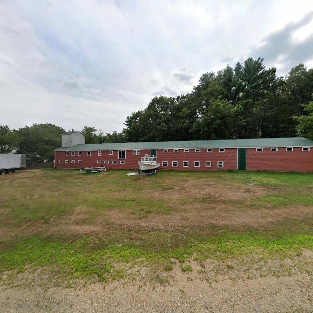 Red barn with green roof and boats parked outside, surrounded by trees and cloudy sky.