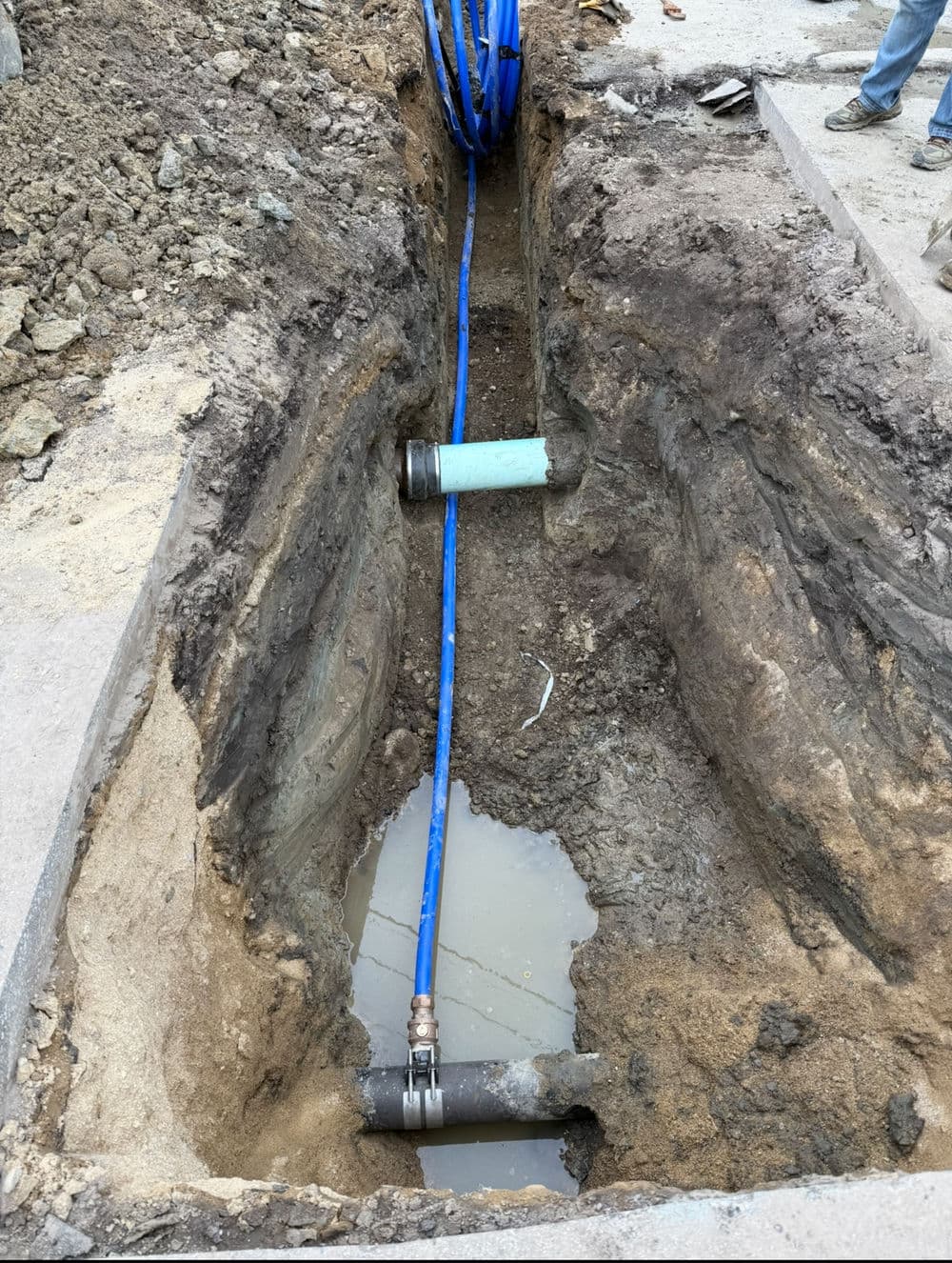 Excavated trench with pipes and blue cabling in construction site, muddy ground visible.