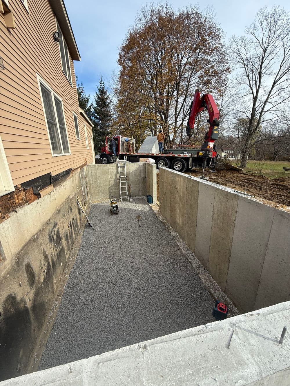 Excavation for a new foundation with gravel, machinery, and a house in the background.