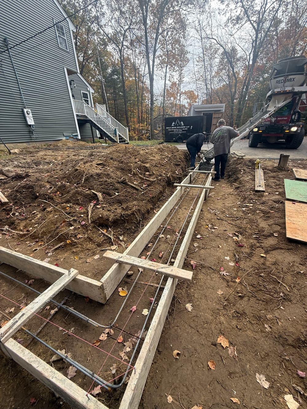 Construction workers installing a concrete foundation with rebar reinforcement in a wooded area.