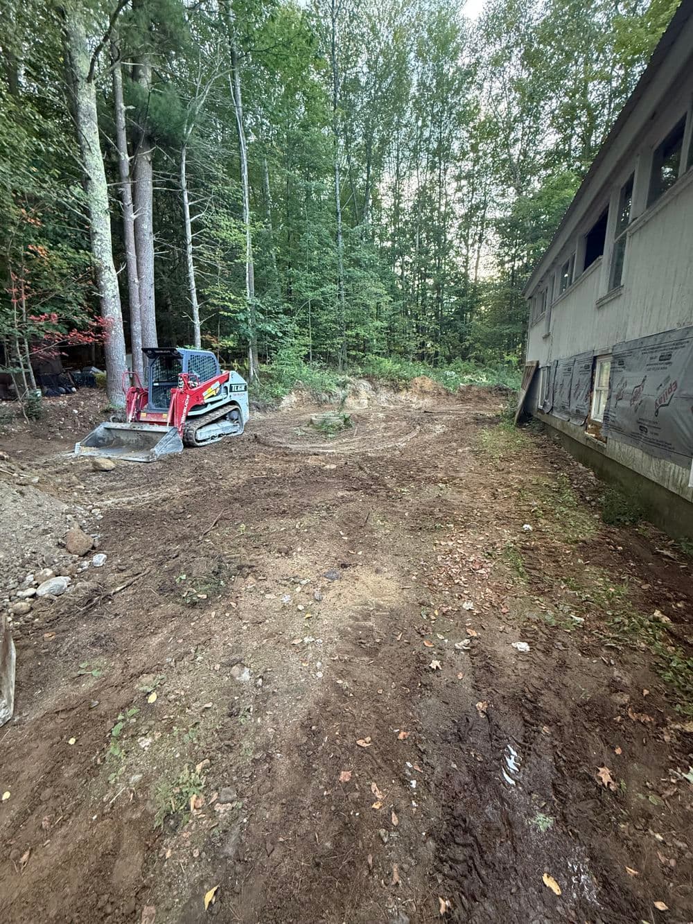 Excavator on cleared land surrounded by trees near a house renovation site.