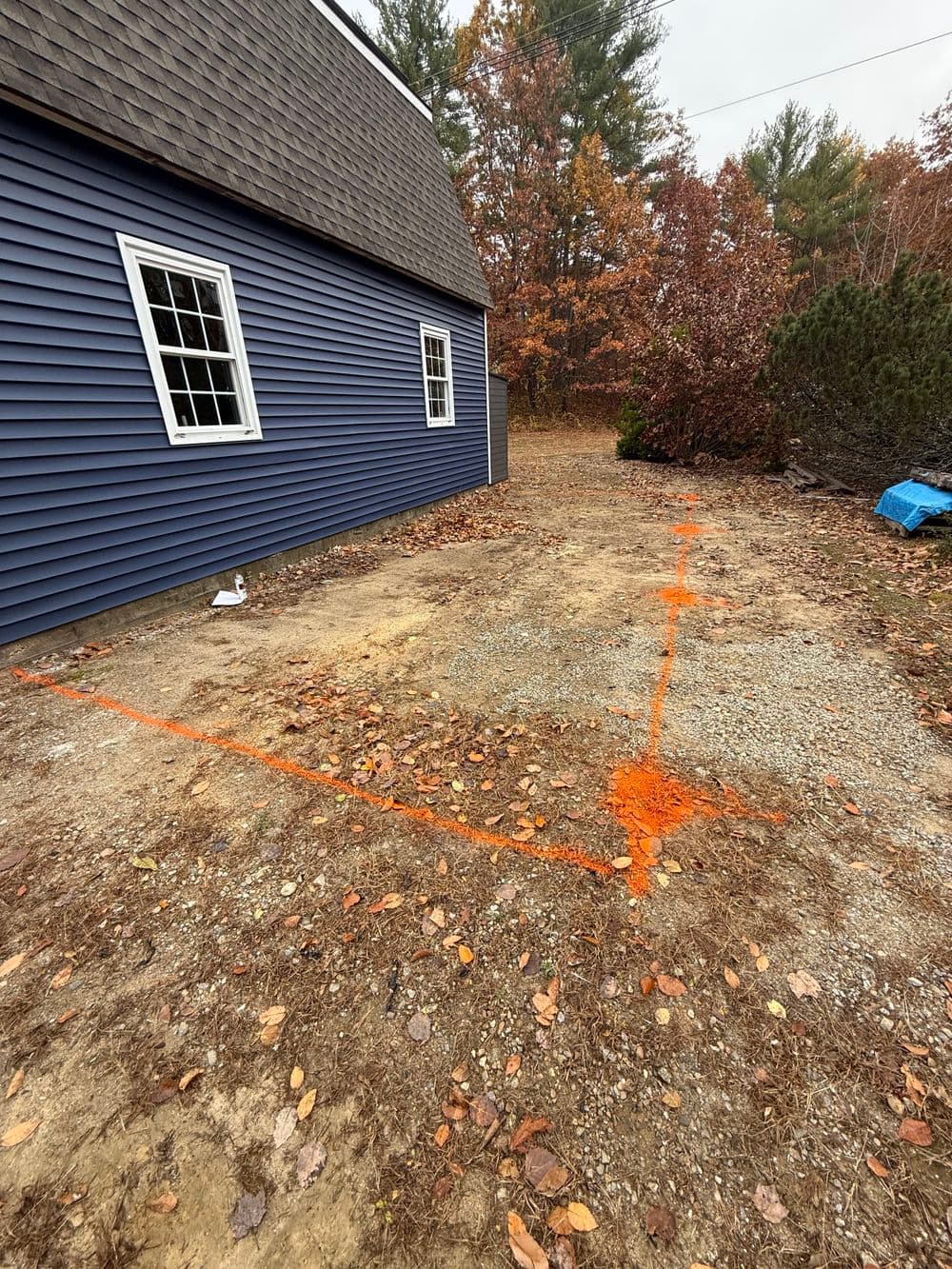 Marked construction area beside blue house with autumn foliage and gravel ground.