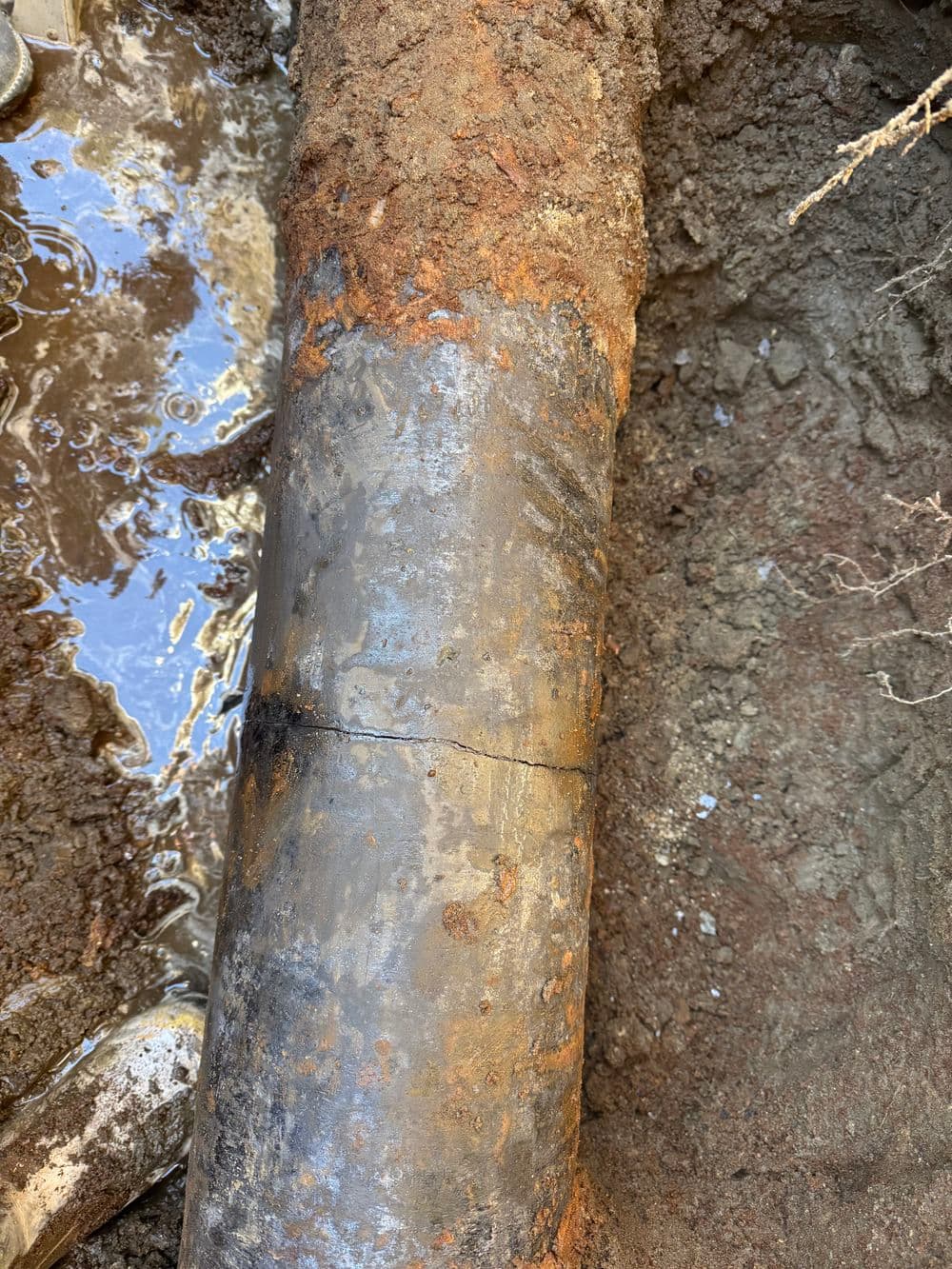 Corroded metal pipe with crack in muddy ground near water source, showing signs of wear.