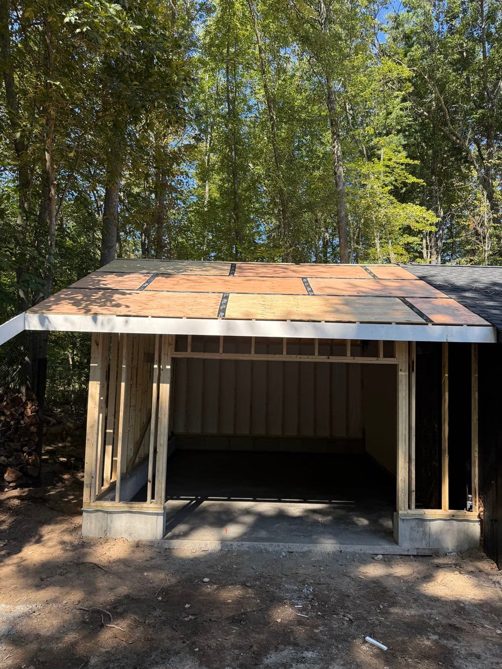 Newly constructed garage with a wooden framework and sloped roof in a wooded area.