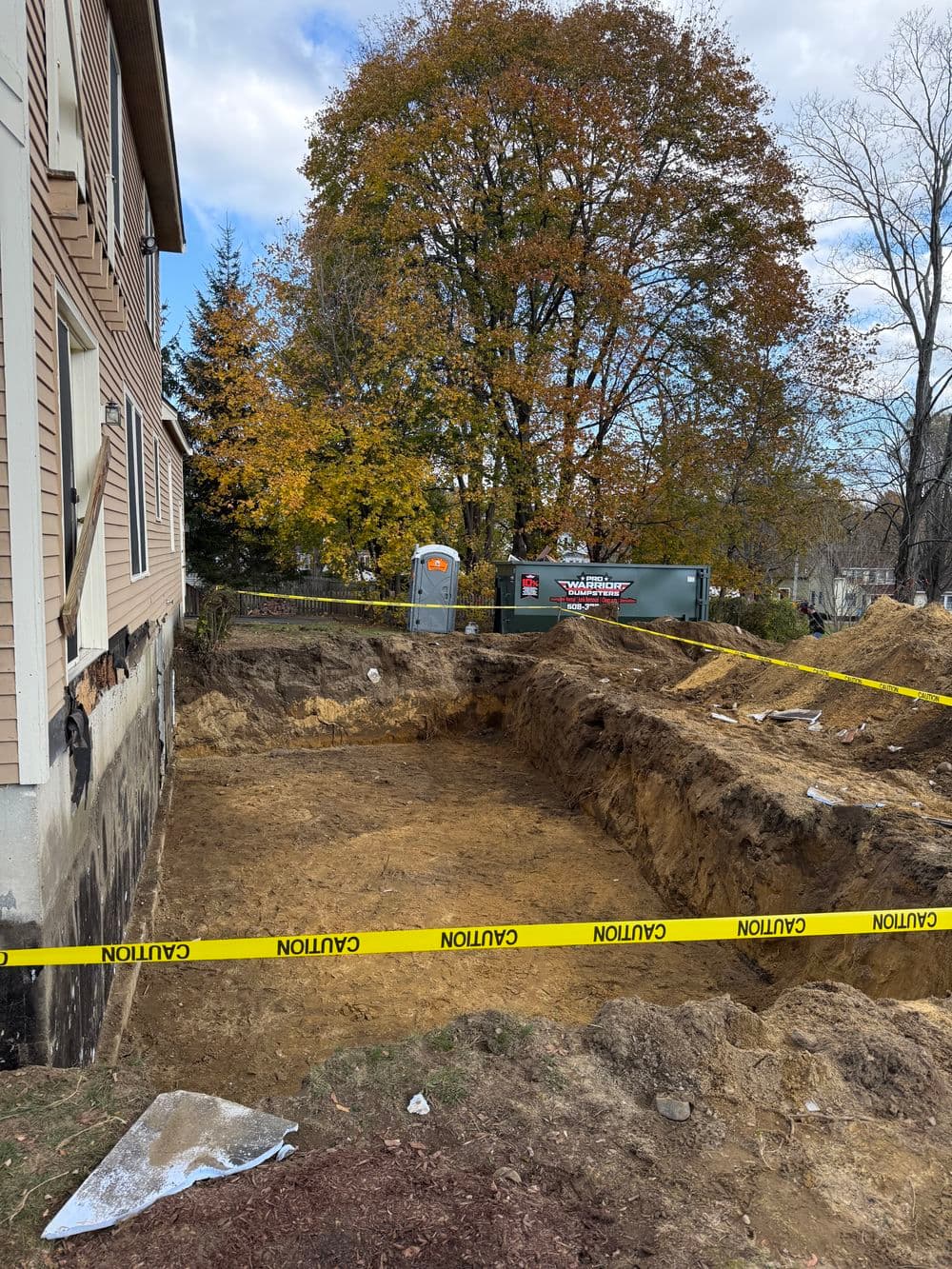 Excavation site with caution tape, portable restroom, and autumn trees in the background.